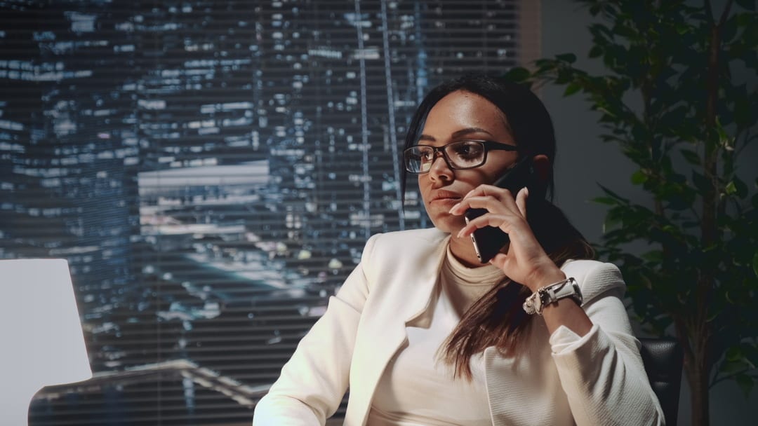 A businesswoman in glasses talks on a phone in an office with a cityscape background at night.