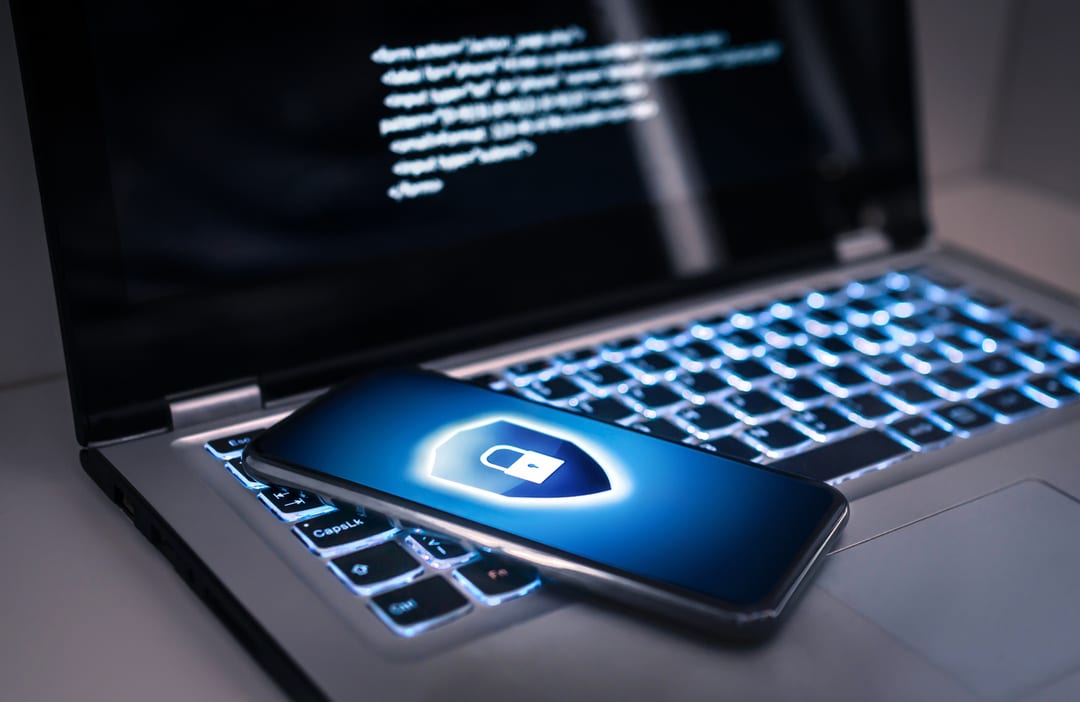 A smartphone with a glowing shield and lock icon rests on an illuminated laptop keyboard.